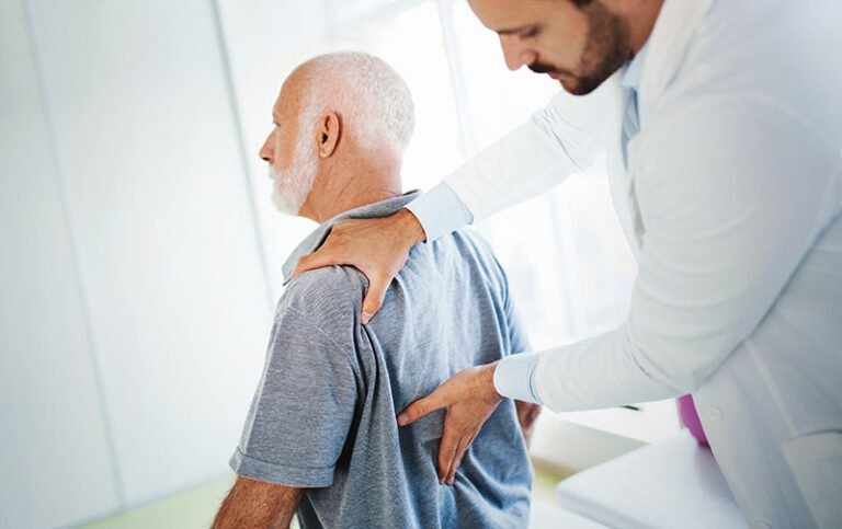 Closeup side view of an early 60's senior gentleman having some lower back pain. He's at doctor's office having medical examination by a male doctor. The doctor is touching the sensitive area and trying to determine the cause of pain.